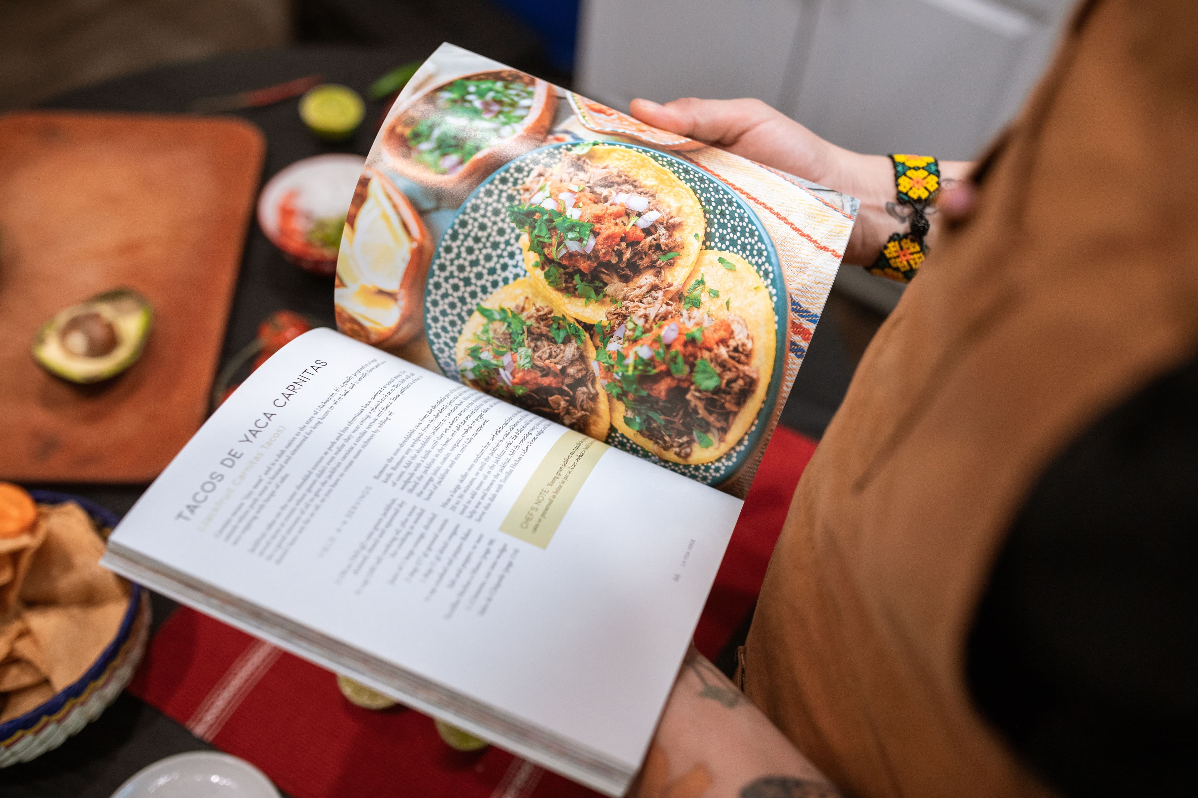 An open cookbook of bright, colourful tacos, held over a laid table